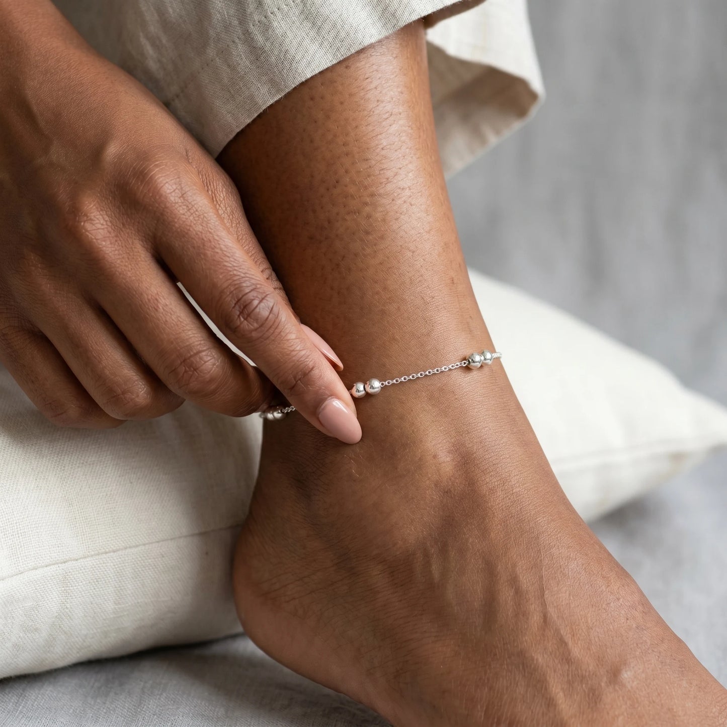 Intimate hand-to-anklet close-up — fingertips touching silver bead station on dainty cable chain