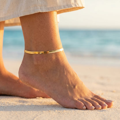 Gold herringbone flat chain anklet worn on tanned ankle on Caribbean beach golden hour