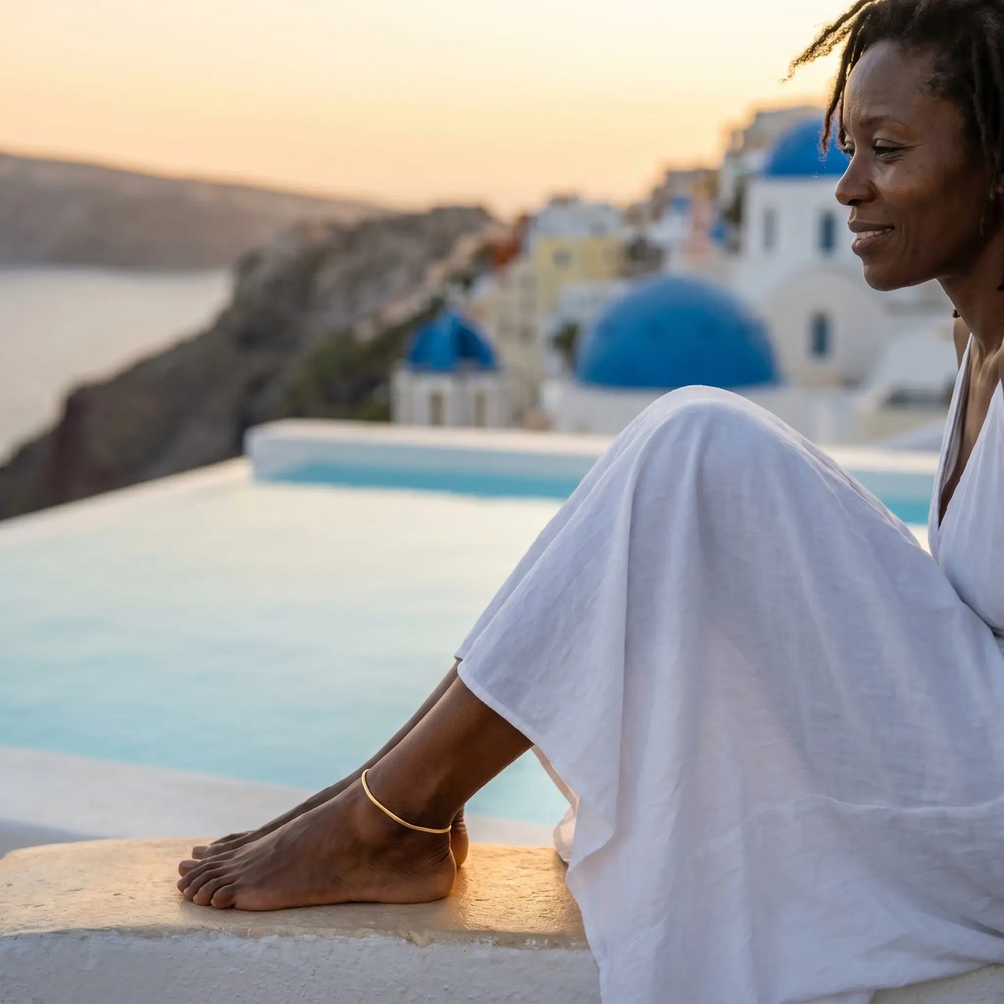 Woman in white maxi dress at Santorini infinity pool wearing gold anklet