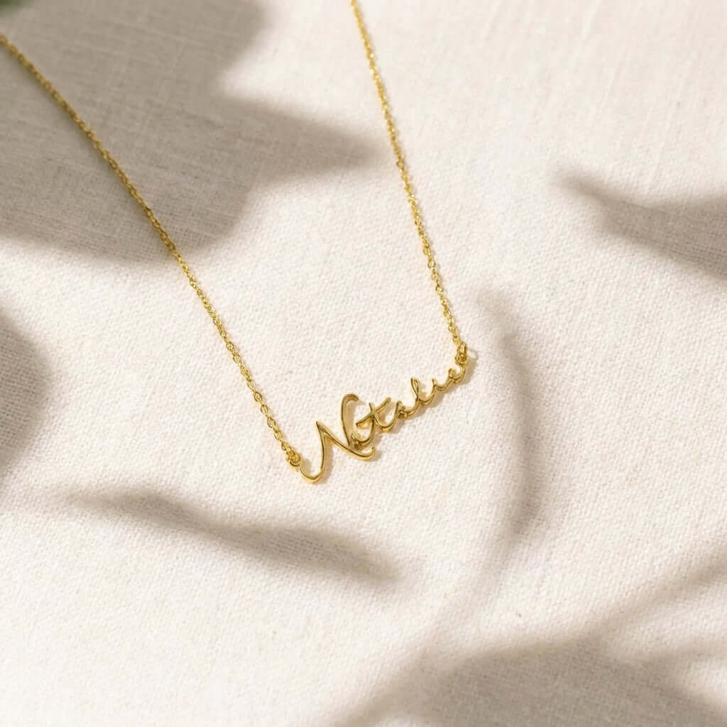 AUREATE Petit Name Necklace featuring elegant gold script on a light background, perfect for personalizing with names.