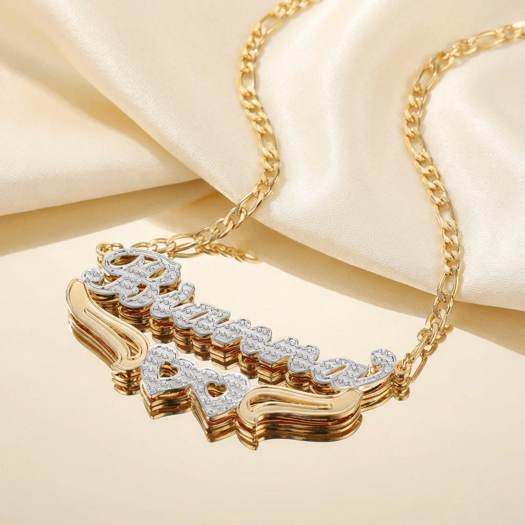 Double Heart Name Necklace with Figaro Chain by AUREATE featuring a gold and silver plated design on a satin background.