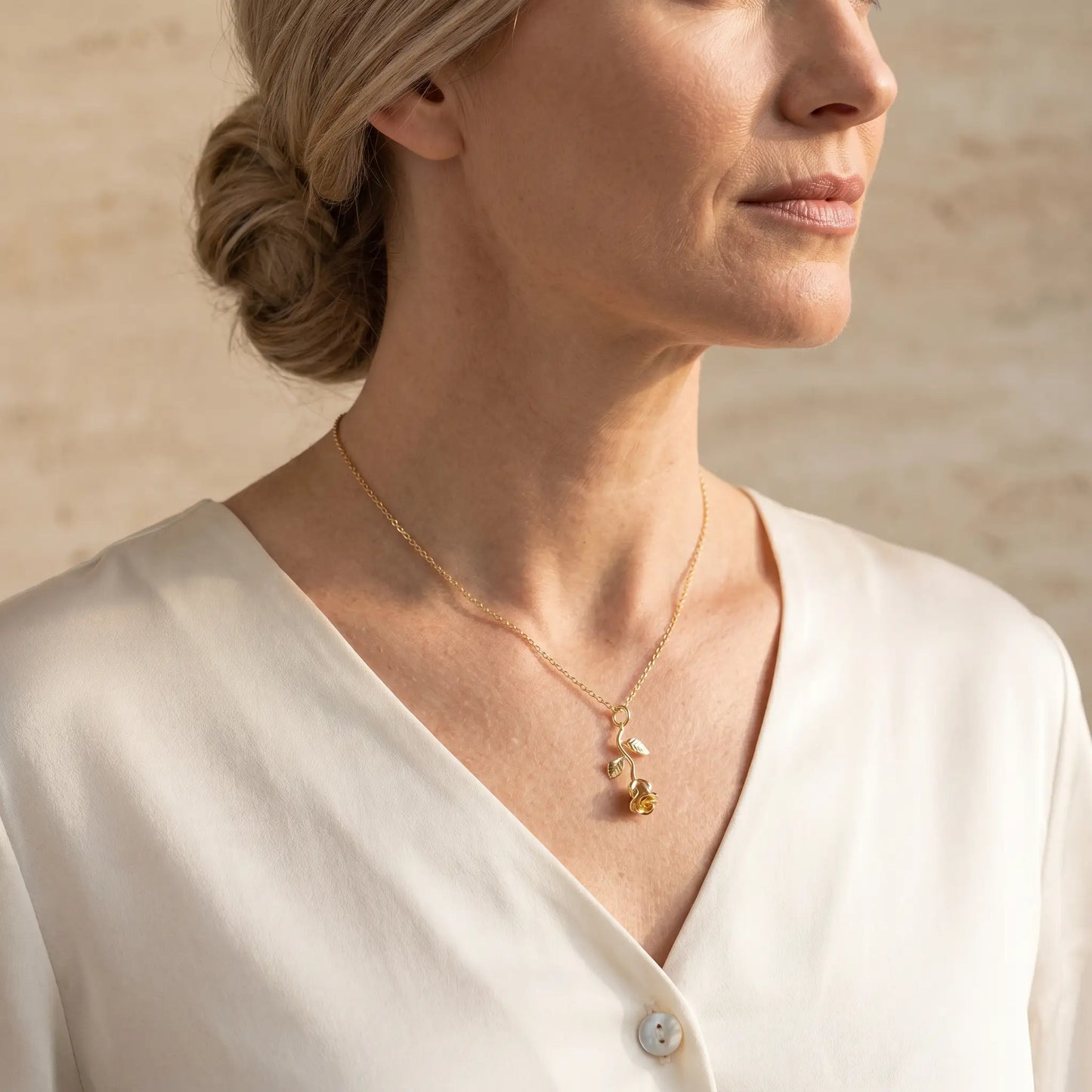 AUREATE Rose Flower Charm Pendant 18K Gold worn 3/4 profile by a mature woman in a cream silk blouse — quiet luxury elegance, warm natural daylight.