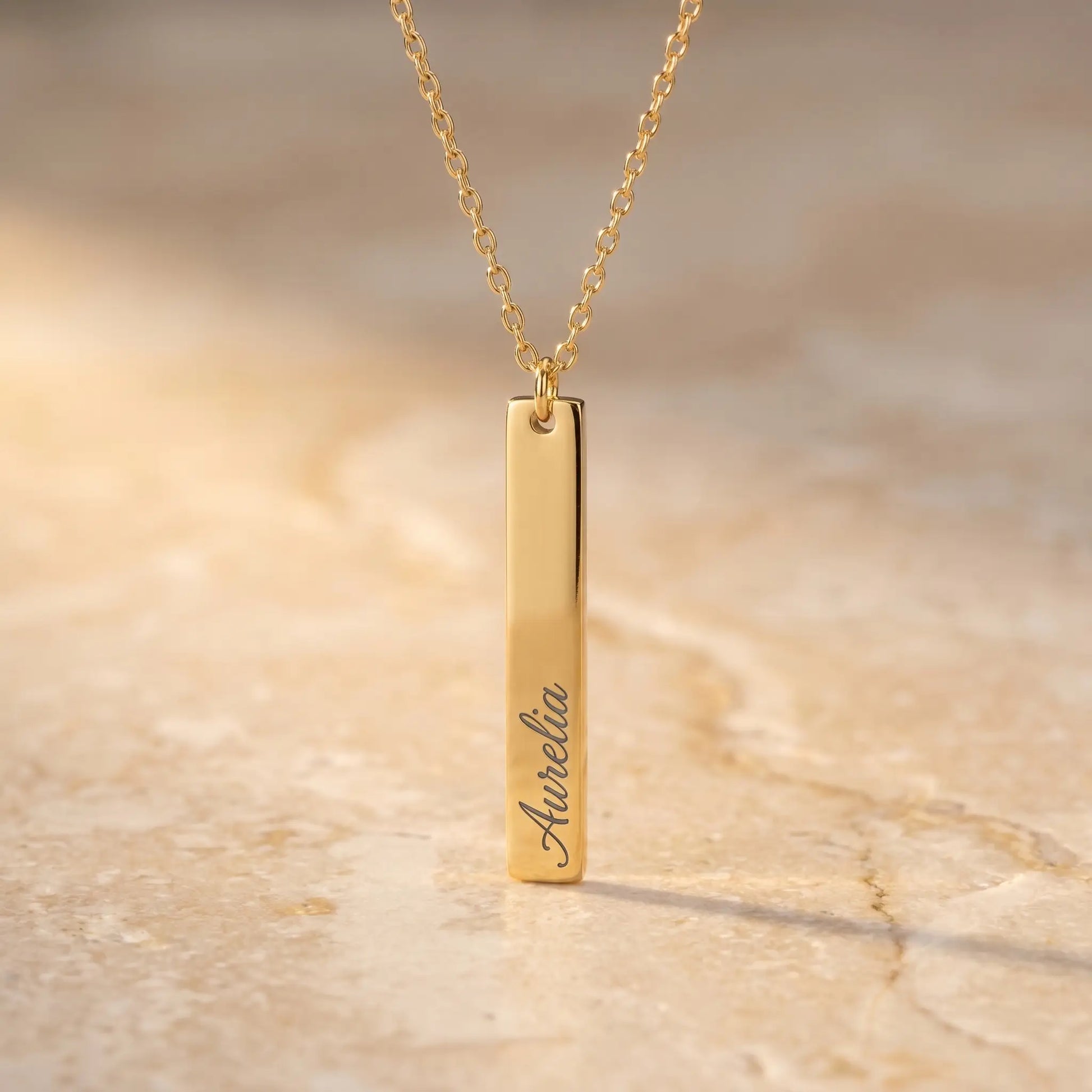 AUREATE Vertical Name Bar Pendant Necklace in 18K gold, engraved with the cursive name Aurelia, displayed on warm travertine background.
