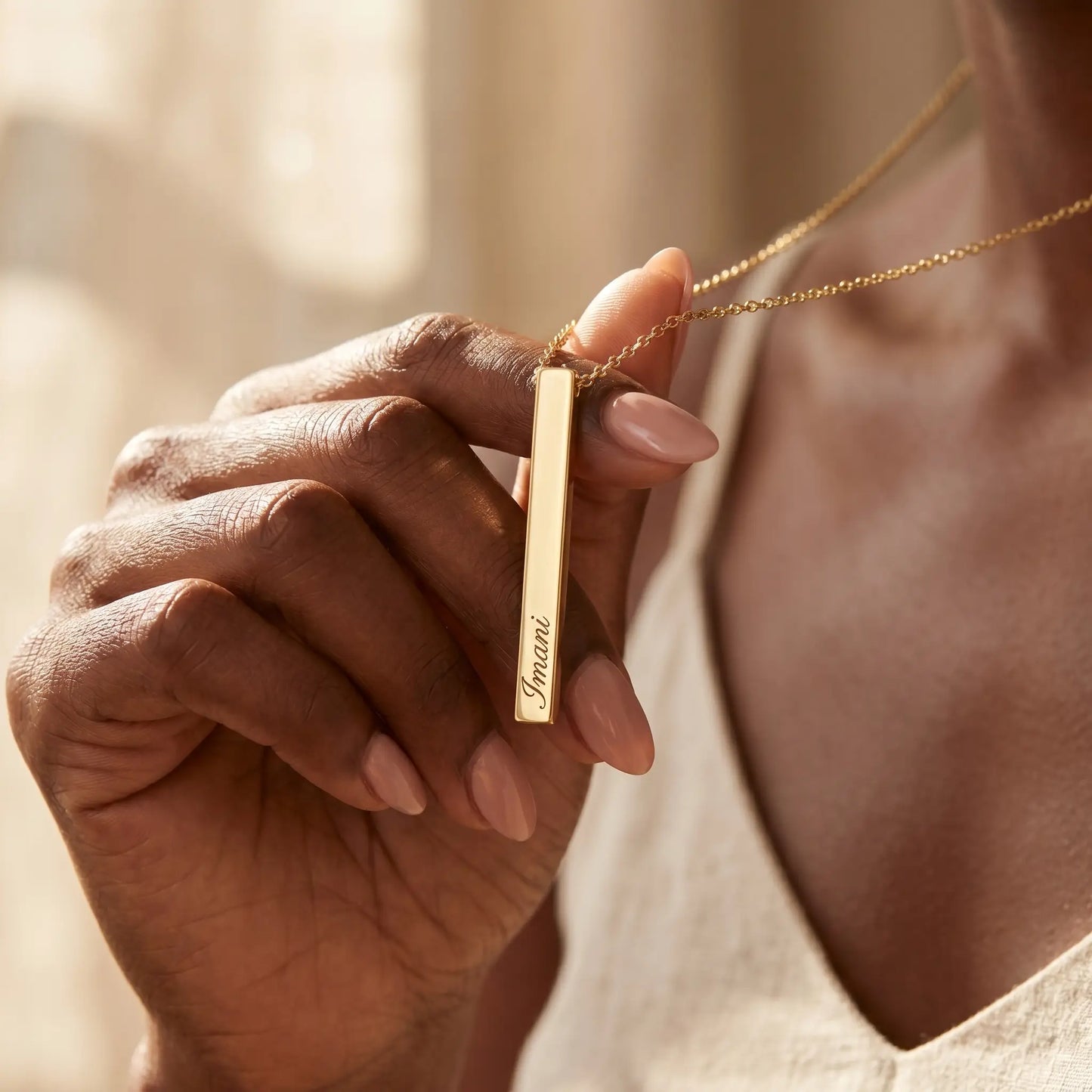 Nigerian woman tenderly holding the AUREATE Vertical Name Bar Pendant in 18K gold engraved Imani, intimate hand-to-jewelry close-up.