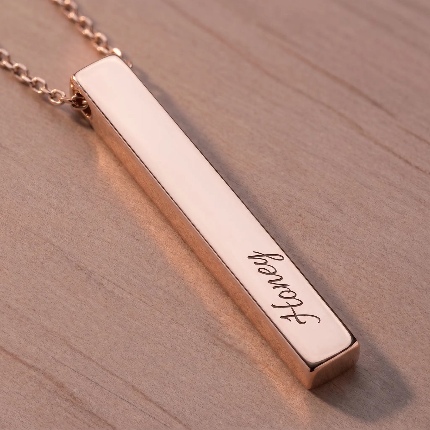 Macro detail of AUREATE Vertical Name Bar Pendant in rose gold engraved cursive Honey, polished pink-gold surface on dusty rose wood.