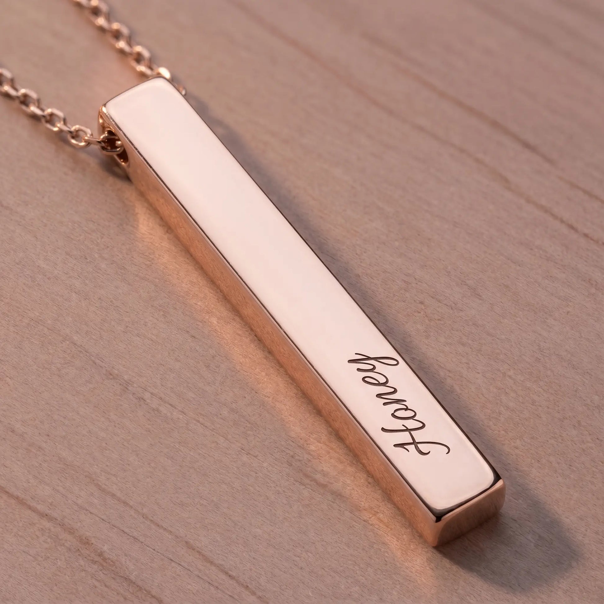 Macro detail of AUREATE Vertical Name Bar Pendant in rose gold engraved cursive Honey, polished pink-gold surface on dusty rose wood.