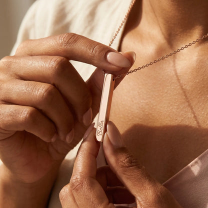 Trinidadian woman tenderly holding the AUREATE Vertical Name Bar Pendant in rose gold engraved Bella, intimate hand-to-jewelry close-up California morning.