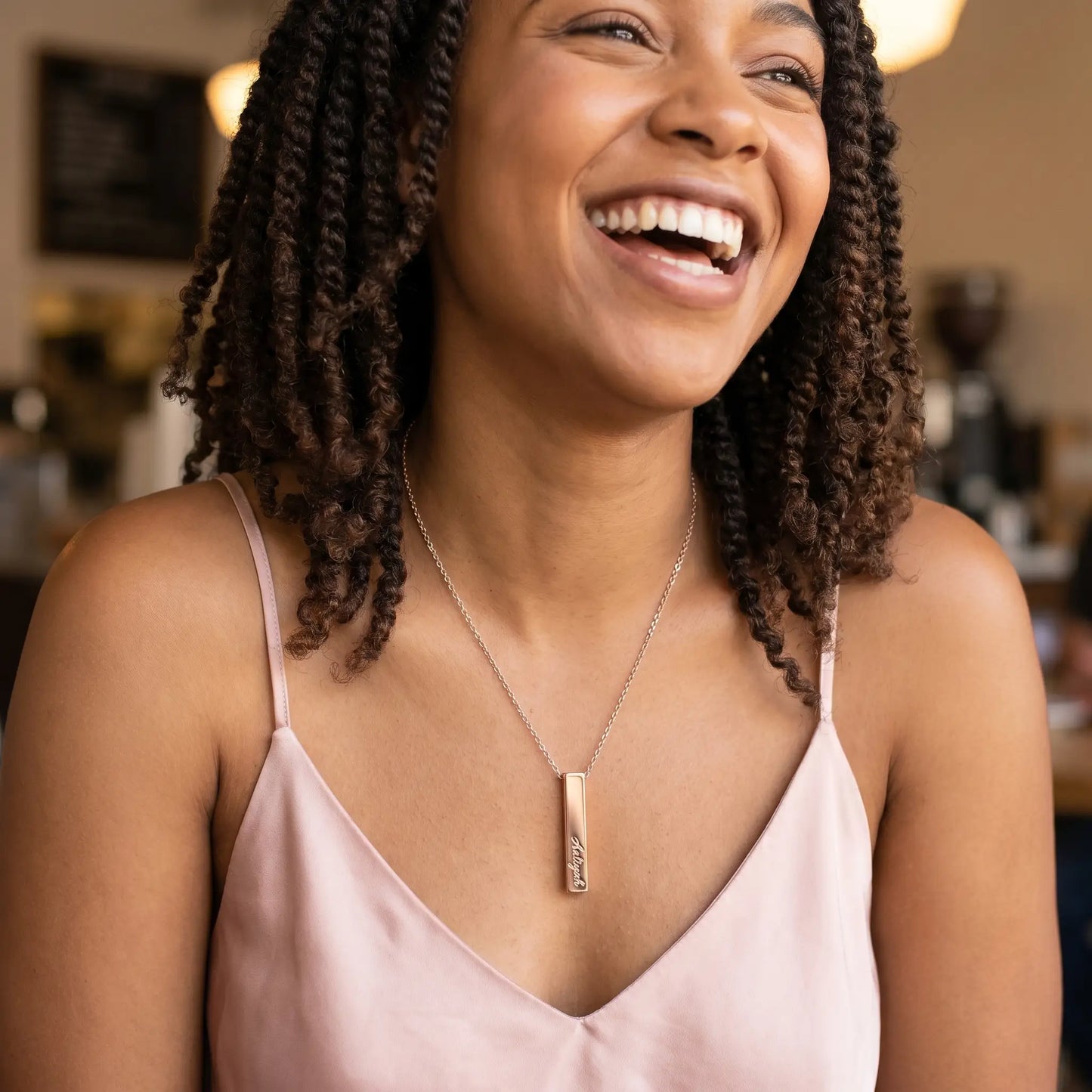 African American woman with twist-out laughing candidly with friend, wearing AUREATE Vertical Name Bar Pendant in rose gold engraved Aaliyah, LA Silver Lake café.