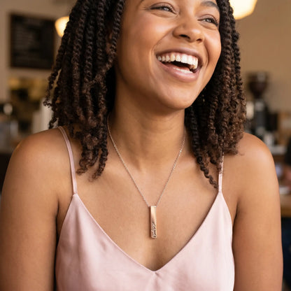African American woman with twist-out laughing candidly with friend, wearing AUREATE Vertical Name Bar Pendant in rose gold engraved Aaliyah, LA Silver Lake café.