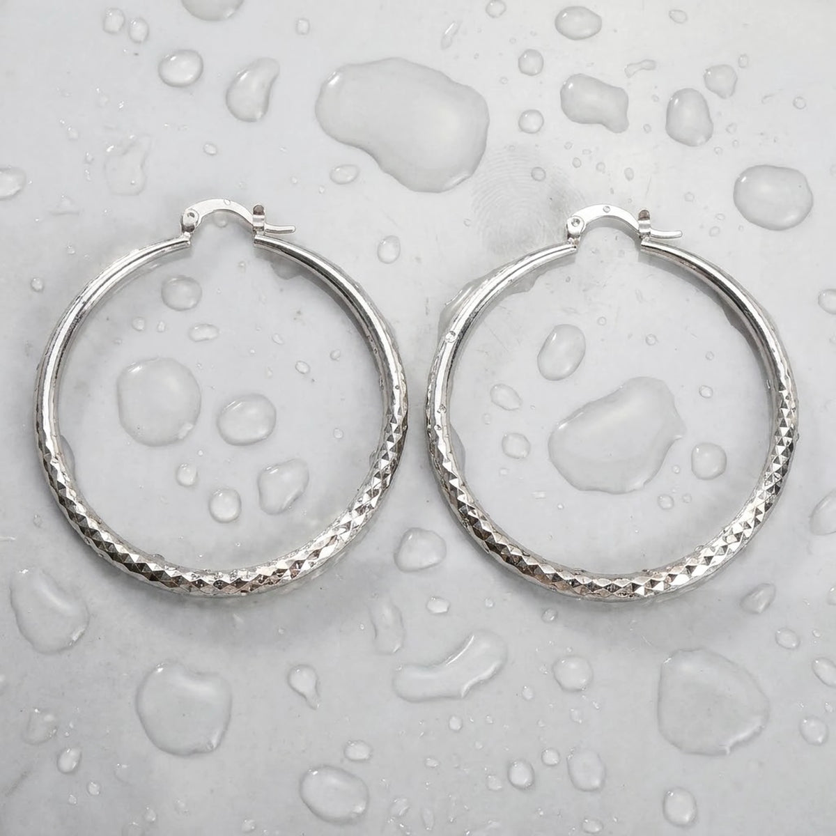 Silver tone stainless steel diamond cut large hoop earrings, still life on a wet surface, waterproof | SKU: AUR-DCTHOP-S