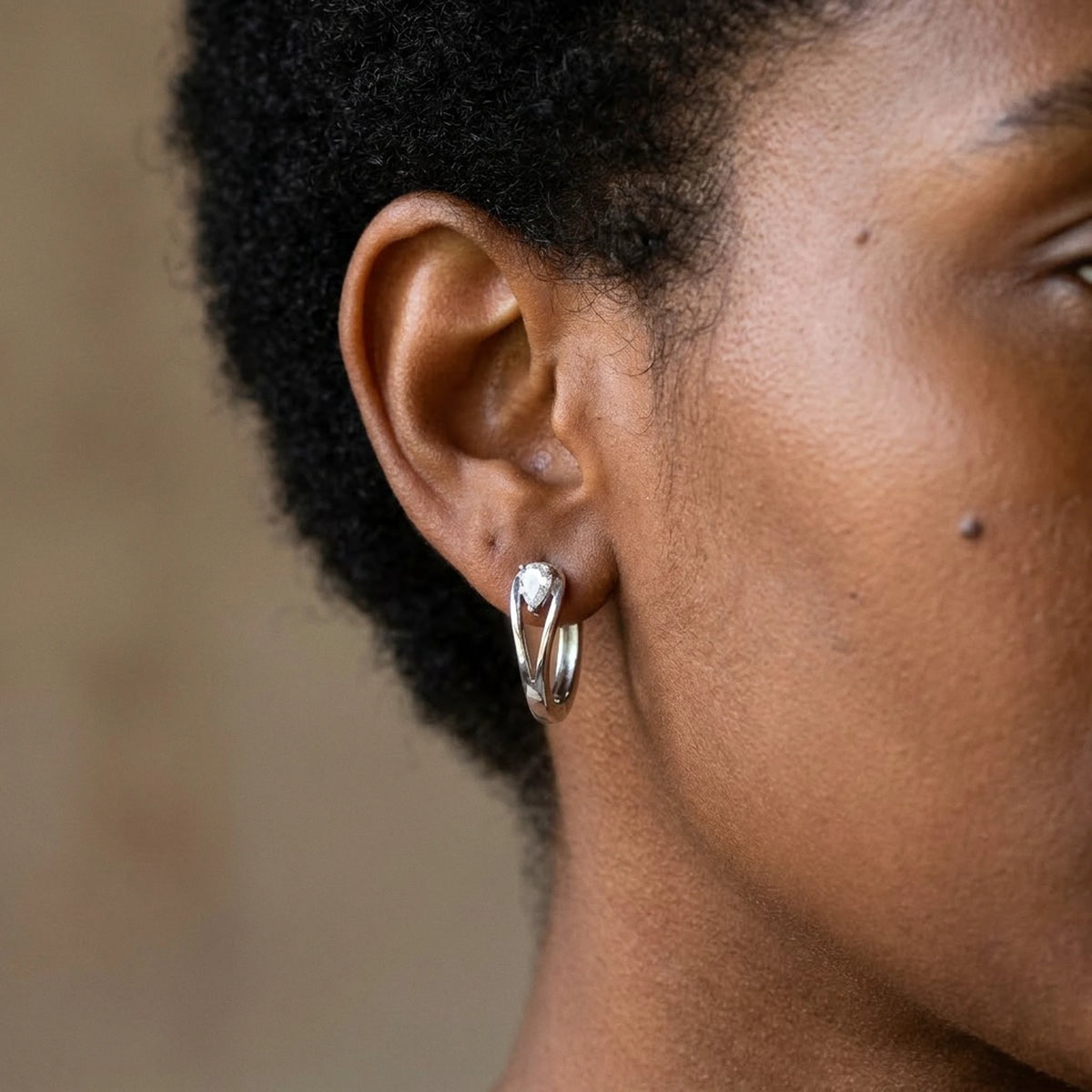 Silver tone stainless steel pear cut crystal huggie hoop earrings, worn close up on the ear