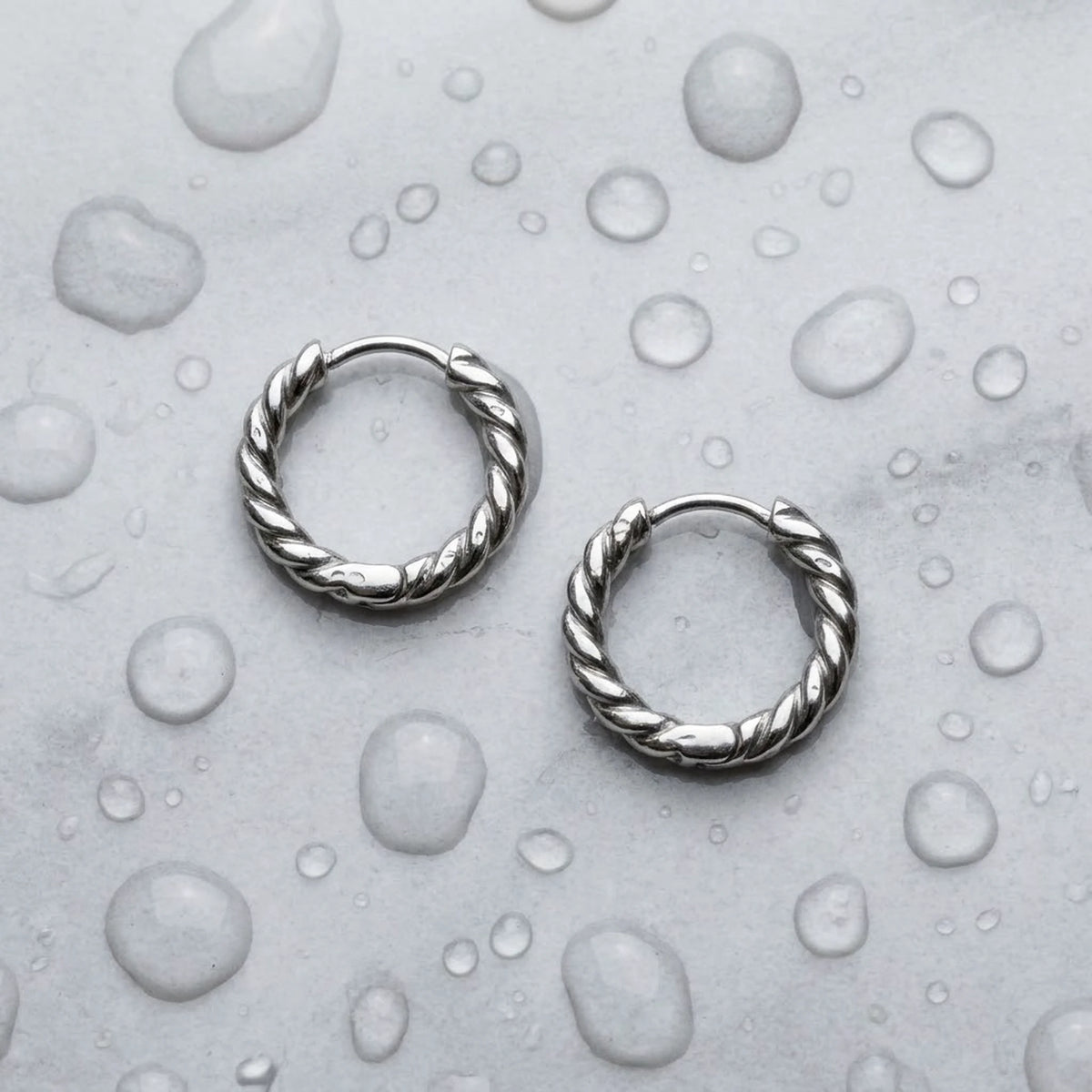 Silver tone stainless steel twisted rope huggie hoop earrings, still life on a wet surface, waterproof | SKU: AUR-TWRHUG-S