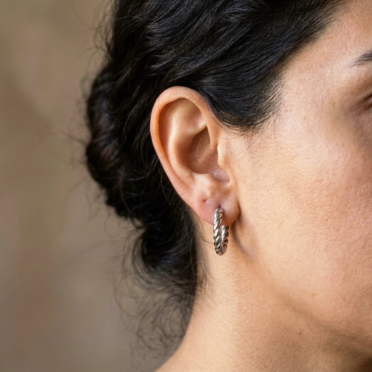 Silver tone stainless steel twisted rope huggie hoop earrings, worn close up on the ear