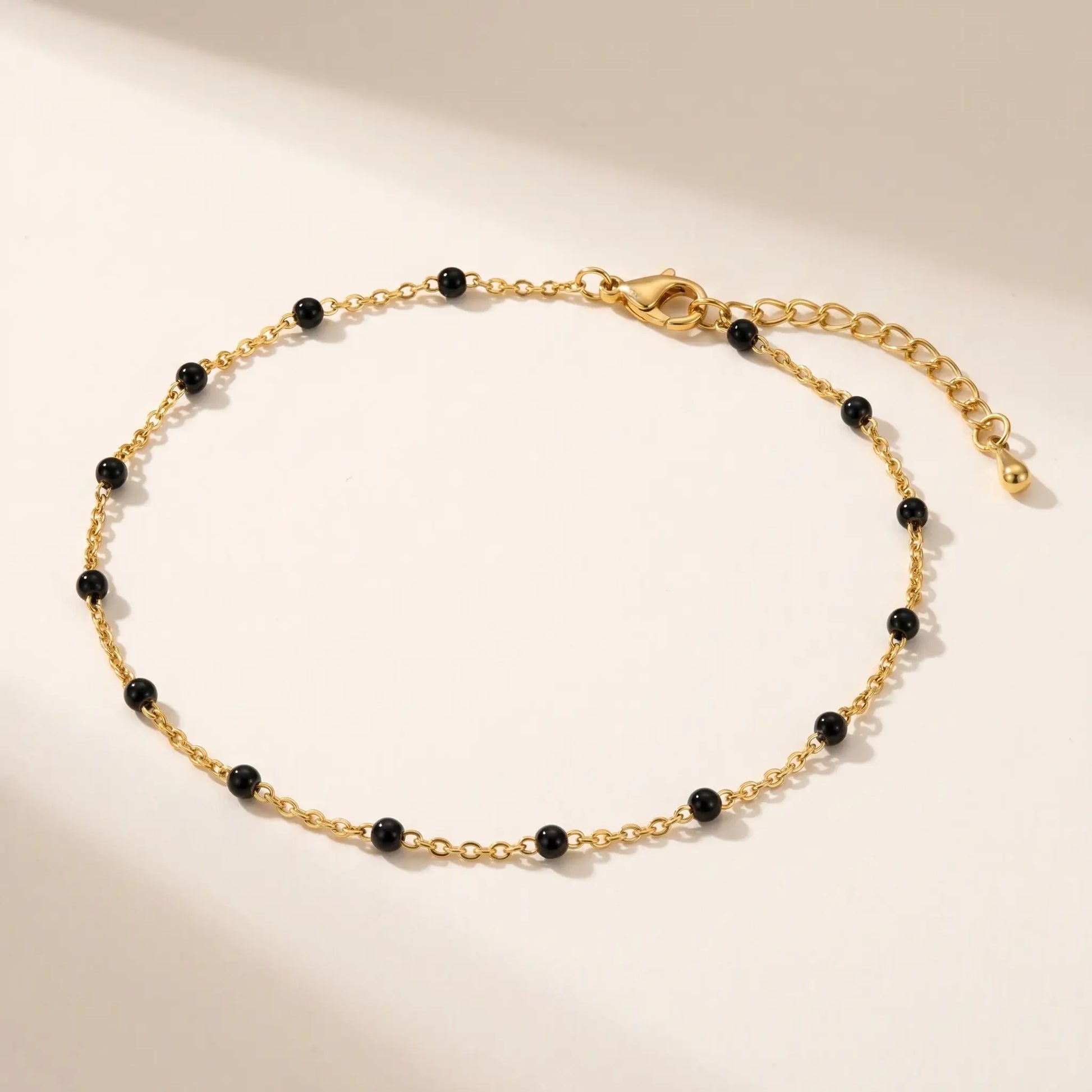 Gold black bead anklet packshot on warm cream background, fine 18K gold cable chain with 12-15 small glossy black enamel beads, lobster clasp and teardrop end charm